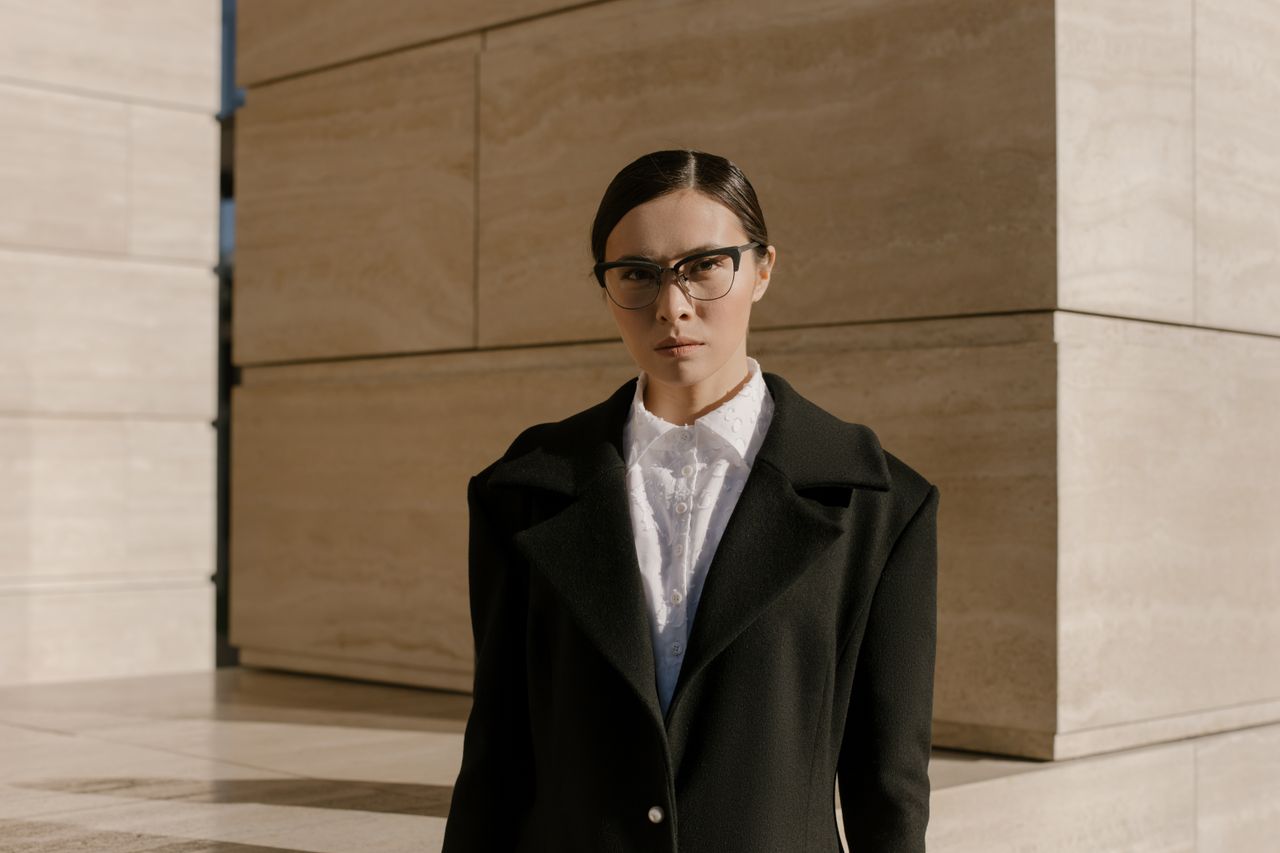 creative-02 Stylish woman in a black coat and eyeglasses posed against a modern building exterior.