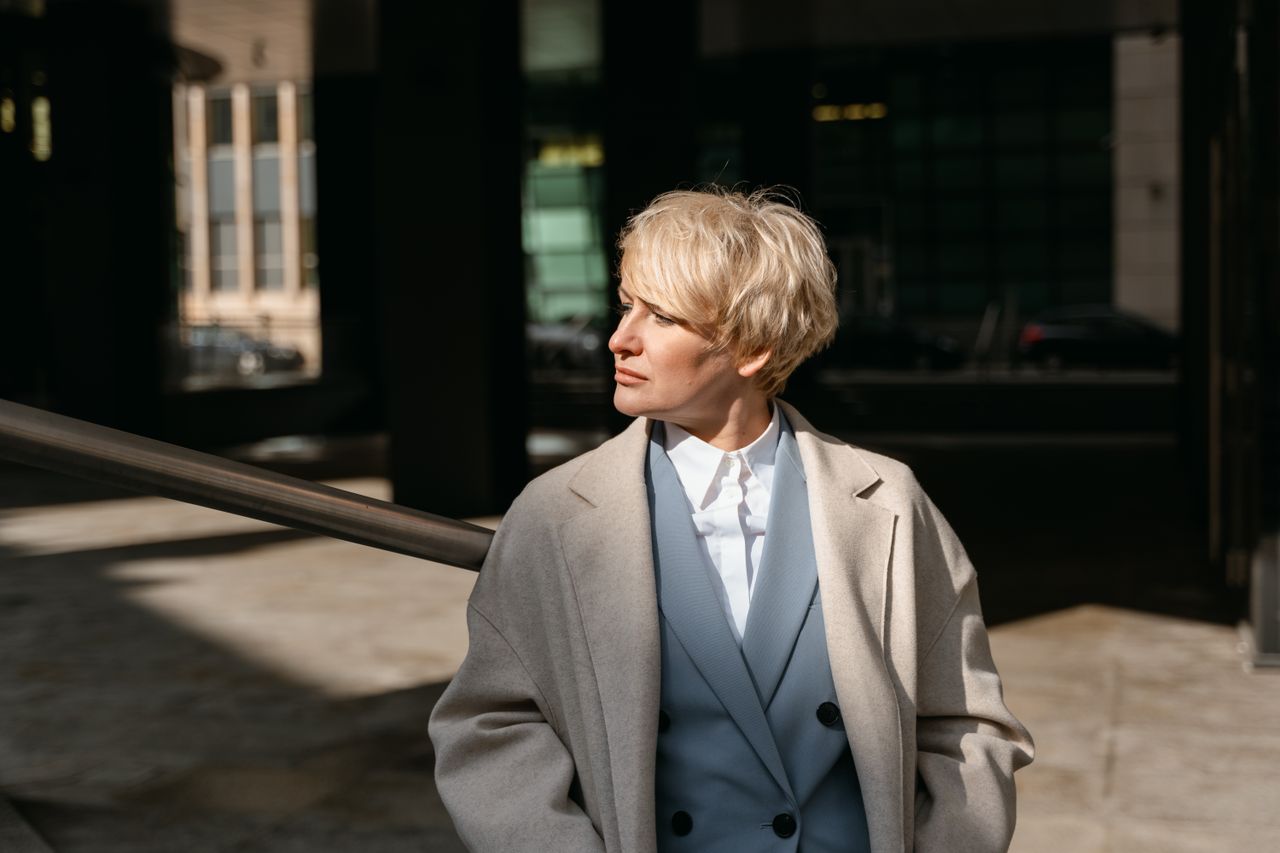 Services-02 Professional blonde woman in a coat, facing away in a sunlit urban environment.
